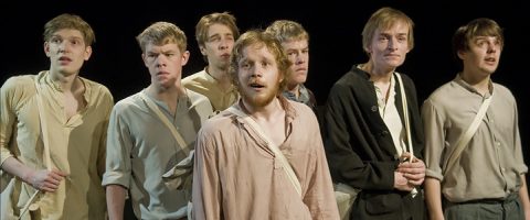 Seitsemän vanhanaikaiseen, yksinkertaiseen vaatetukseen pukeutunutta nuorta miestä seisoo lähekkäin lavalla ja katsoo huolestuneina ja intensiivisin ilmein eteenpäin, ikään kuin reagoisi johonkin kameran ulkopuolella tapahtuvaan.