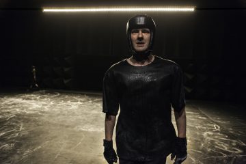 Mustaan kypärään, hanskoihin ja tummaan t-paitaan pukeutunut mies seisoo hämärässä huoneessa, jonka lattiat ovat rapautuneet ja jonka takana on rivistö valoja. Tausta on tumma ja hieman epätarkka.