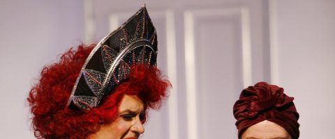 Kaksi henkilöä, joilla on ylleen taidokkaat puvut, mukaan lukien dramaattiset päähineet ja peruukit. Toisella on suuri punainen peruukki ja teräväkärkinen hattu ja toisella syvänpunainen turbaani. Molemmat seisovat vaalean taustan edessä.