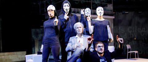 Kuusi ihmistä poseeraa lavalla; neljä seisoo takana mustissa vaatteissa ja Guy Fawkes -naamareissa, kun taas kaksi istuu edessä, toinen harmaassa puvussa ja valkoisessa peruukissa ja toinen mustissa silmälaseissa. Asetelma on teatraalinen ja dramaattinen.