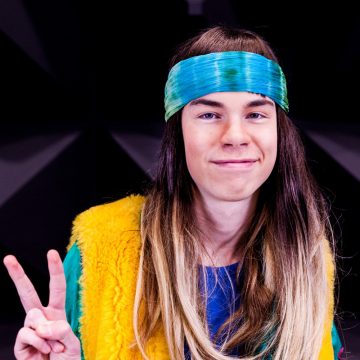 Hymyilevä pitkähiuksinen henkilö, jolla on sininen ja vihreä otsanauha, keltainen karvalakki ja sininen paita, pitää toisella kädellään rauhanmerkkiä tummalla taustalla.