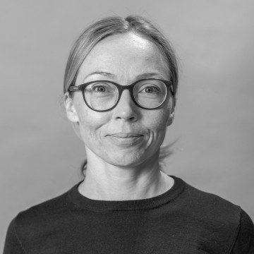 Mustavalkoinen muotokuva naisesta, jolla on vaaleat taakse vedetyt hiukset, silmälasit ja tumma toppi, hymyilee hieman ja katsoo suoraan kameraan tavallista taustaa vasten.