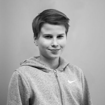 Lyhythiuksinen nuori henkilö, jolla on vetoketjullinen Nike-huppari, seisoo vaalean taustan edessä hymyillen pehmeästi. Kuva on mustavalkoinen.