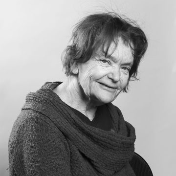 Vanhemmalla naisella, jolla on lyhyet, sotkuiset hiukset, on kuvioitu villapaita ja hän hymyilee lempeästi kameralle tässä mustavalkoisessa muotokuvassa tavallista taustaa vasten.