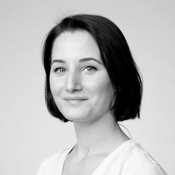 Mustavalkoinen muotokuva naisesta, jolla on suorat tummat polkkatukkaiset hiukset, päällään vaalea toppi, katselee kameraan hymyillen tavallista taustaa vasten.
