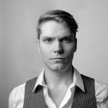 Nuori mies, jolla on lyhyet, siististi laitetut hiukset, tuijottaa vakavana kameraan. Hänellä on yllään valkoinen paita, jonka kaulus on auki, ja tumma, nastaraitainen liivi. Tausta on tavallinen ja vaalea. Kuva on mustavalkoinen.