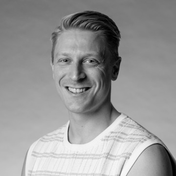 Hymyilevä mies, jolla on lyhyet, vaaleat hiukset, pukeutuu hihaton, vaaleaan, kuvioituun paitaan. Kuva on mustavalkoinen ja siinä on yksinkertainen, sumea tausta.