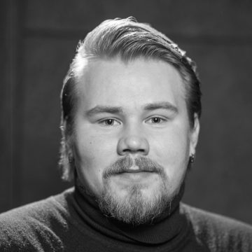 Mustavalkoinen muotokuva henkilöstä, jolla on vaalea iho, lyhyet liukuvat hiukset, parta ja viikset, päällään tumma villapaita ja pienet korvakorut, katsoo suoraan kameraan neutraali ilme.