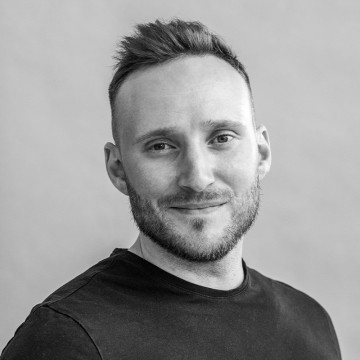 Lyhyet ruskeat hiukset ja partainen mies, jolla on yllään musta t-paita, hymyilee lempeästi ja katsoo kameraan vaaleaa taustaa vasten.