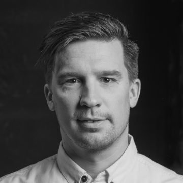 Mustavalkoinen muotokuva miehestä, jolla on lyhyet, siististi muotoillut hiukset, päällään napitettu vaalea paita, katsoo suoraan kameraan neutraali ilme.