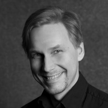 Mustavalkoinen muotokuva vaaleahiuksisesta miehestä, jolla on lyhyt parta ja viikset ja joka hymyilee kameralle. Hänellä on tumma kauluspaita ja hän poseeraa tavallisen, kuvioidun taustan edessä.