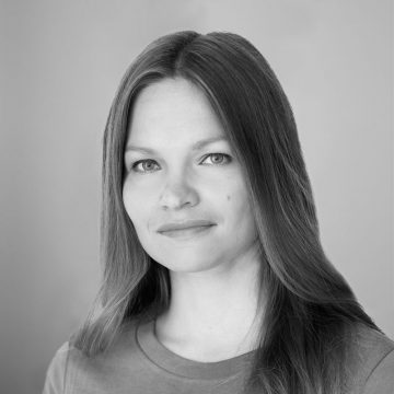 Nainen, jolla on pitkät suorat hiukset ja vaaleat silmät, katsoo kameraan neutraalilla ilmeellä. Hänellä on yllään yksinkertainen paita, ja tausta on yksinkertainen ja vaalea. Kuva on mustavalkoinen.