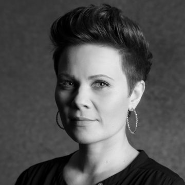 Nainen, jolla on lyhyet, muotoillut hiukset ja korvarenkaat, katsoo kameraan neutraalilla ilmeellä. Kuva on mustavalkoinen ja siinä on pehmeä, kuvioitu tausta. Naisella on yllään tumma toppi.