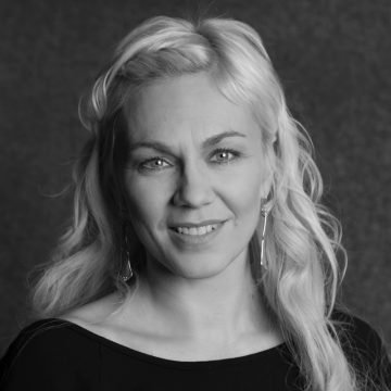 Nainen, jolla on pitkät, vaaleat, aaltoilevat hiukset ja vaaleat silmät, hymyilee pehmeästi. Hänellä on roikkuvat korvakorut ja tumma toppi, ja hän poseeraa tummaa, kuvioitua taustaa vasten. Kuva on mustavalkoinen.