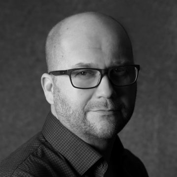 Kaljupäinen mies, jolla on tummat suorakulmaiset silmälasit ja ruutupaita, katsoo kameraan neutraalilla ilmeellä. Kuva on mustavalkoinen ja siinä on tavallinen, kuvioitu tausta.