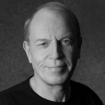 Keski-ikäinen henkilö, jolla on lyhyet vaaleat hiukset, hymyilee pehmeästi kameralle. Hänellä on yllään tumma paita, ja hänet näytetään mustavalkoista, tummaa taustaa vasten.