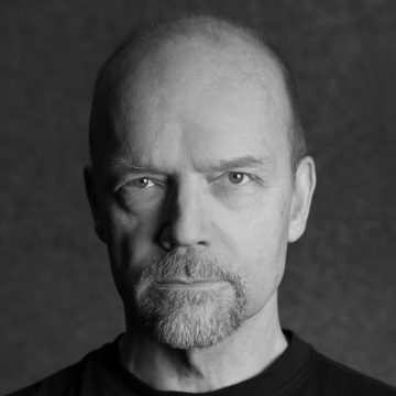 Keski-ikäinen mies, jolla on kalju pää, vaalea iho ja lyhyt parta, katsoo suoraan kameraan vakavalla ilmeellä. Tausta on tavallinen ja tumma, ja kuva on mustavalkoinen.