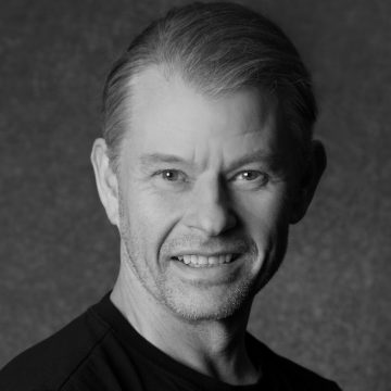 Mustavalkoinen muotokuva keski-ikäisestä, lyhyet vaaleat hiukset omaavasta miehestä, joka hymyilee ja katsoo kameraan. Hänellä on yllään tumma t-paita, ja tausta on yksivärinen ja kuvioitu.