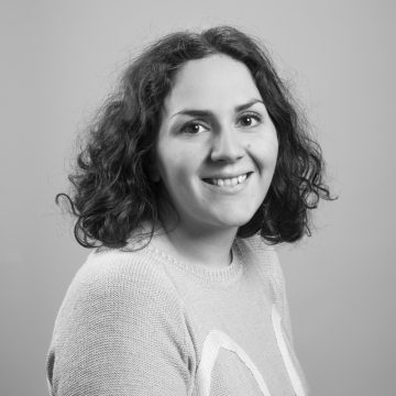 Kiharatukkainen nainen hymyilee kameralle. Hänellä on yllään vaalea villapaita ja hän poseeraa vaaleanharmaata taustaa vasten. Kuva on mustavalkoinen.