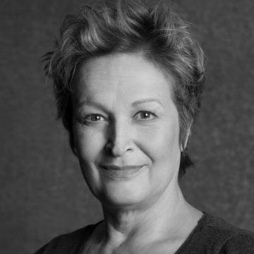 Mustavalkoinen muotokuva vanhemmasta naisesta, jolla on lyhyet, sekaiset hiukset ja joka hymyilee pehmeästi kameralle yksinkertaista, kuvioitua taustaa vasten. Hänellä on yllään yksinkertainen toppi.