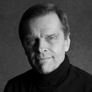Mustavalkoinen muotokuva vanhemmasta lyhythiuksisesta miehestä, jolla on tumma poolopaita ja joka katsoo suoraan kameraan neutraalilla ilmeellä tavallista taustaa vasten.