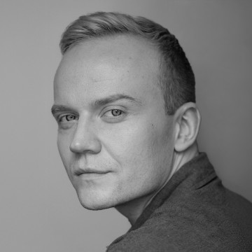 Lyhyet vaaleat hiukset ja vaaleat silmät omaava mies katsoo olkansa yli kohti kameraa. Muotokuva on mustavalkoinen, pehmeä valaistus korostaa miehen kasvonpiirteitä ja tausta on neutraali.