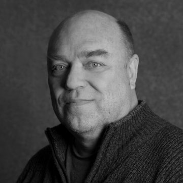 Keski-ikäinen mies, jolla on lyhyet hiukset ja hento hymy, on pukeutunut vetoketjulliseen villapaitaan. Kuva on mustavalkoinen ja siinä on kuvioitu tausta.