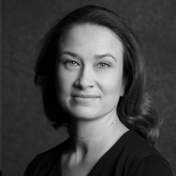 Mustavalkoinen muotokuva naisesta, jolla on hartioiden pituiset aaltoilevat hiukset, yllään tumma toppi, katselee kameraan hymyillen tummaa taustaa vasten.