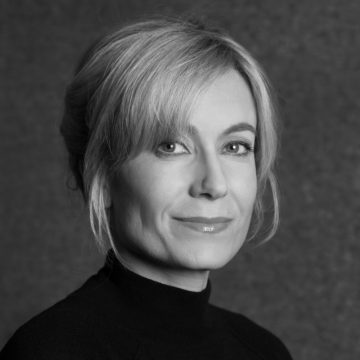 Mustavalkoinen muotokuva naisesta, jolla on vaaleat taaksepäin muotoillut hiukset, yllään tumma korkeakaulainen toppi, hymyilee hieman ja katsoo kameraan yksiväristä taustaa vasten.