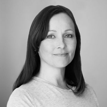 Nainen, jolla on pitkät tummat hiukset ja vaaleat silmät, hymyilee pehmeästi. Hänellä on yllään vaalea toppi ja hän poseeraa mustavalkoisessa muotokuvassa vaaleanharmaata taustaa vasten.