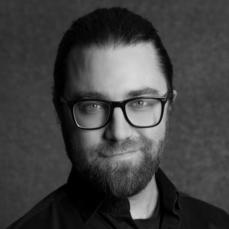 Mustavalkoinen muotokuva parrakas mies, jolla on silmälasit ja taakse vedetyt pitkät hiukset, yllään tumma paita, katselee kameraan hymyillen, kuvioitu tausta.