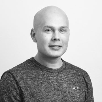 Kaljupäinen mies, jolla on tumma, kuvioitu pusero, poseeraa vaalean taustan edessä. Hän on kameraan päin ja hänellä on neutraali ilme. Kuva on mustavalkoinen.