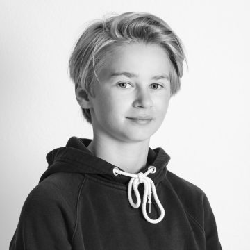Mustavalkoinen muotokuva vaaleahiuksisesta nuoresta henkilöstä, jolla on huppari päällään ja joka katsoo kameraan neutraali ilme kasvoillaan tavallista taustaa vasten.