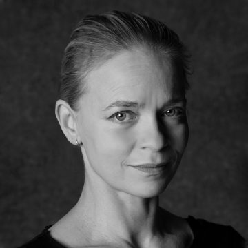 Mustavalkoinen muotokuva naisesta, jolla on taaksepäin vedetyt vaaleat hiukset ja tumma toppi, joka katsoo kameraan hymyillen, tummaa taustaa vasten.
