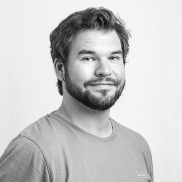 Mustavalkoinen muotokuva miehestä, jolla on lyhyet, aaltoilevat hiukset ja parta, yllään rento T-paita. Hän katsoo hieman sivulle ja hymyilee pehmeästi, ja hänen takanaan on neutraali tausta.