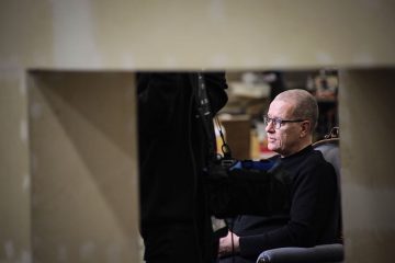 Silmälasipäinen mies, jolla on musta villapaita, istuu tuolissa, joka näkyy suorakulmaisen aukon läpi. Tausta on epätarkka, ja etualalla näkyy osittain toinen henkilö.
