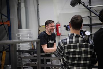 Mies hymyilee ja puhuu teollisuusympäristössä kameramiehen kuvatessa. Miehellä on musta t-paita ja hän seisoo metallirakenteiden lähellä; taustalla näkyy valoja, hyllyjä ja sammutin.