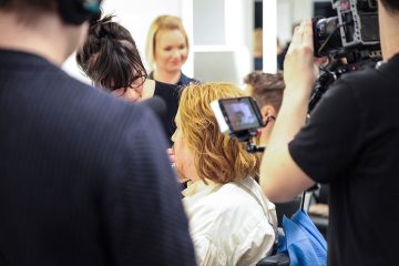 Vaaleanruskeat hiukset omaava henkilö istuu, kun meikkaaja meikkaa. Useat kuvausryhmän jäsenet ja kamera kuvaavat kohtausta kirkkaasti valaistussa studiossa.