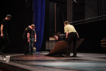 Neljä näyttämöhenkilökunnan jäsentä nostaa ja asettaa suuren puisen laudan hämärästi valaistulle teatterin lavalle, jonka taustalla näkyvät siniset verhot ja lavastemateriaalit.