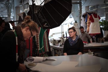 Useat ihmiset työskentelevät muotisuunnittelustudiossa ompelukoneiden, kankaan ja pukumuottien parissa. Nainen istuu pöydän ääressä, kun toinen seisoo ja mittaa materiaalia. Suuret valot roikkuvat yläpuolella, ja taustalla roikkuu vaatteita.