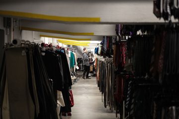 Henkilö seisoo hämärässä varastohuoneessa tai kellarissa vaatehyllyjen rivien välissä, roikkuvien vaatteiden ja asusteiden ympäröimänä.