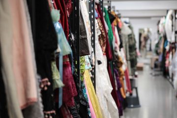 Värikkäitä pukuja ja vaatteita roikkuu riveittäin telineissä hyvin valaistussa varasto- tai pukuhuoneessa, ja telineiden välissä on pitkä käytävä. Kohtaus viittaa teatterin tai elokuvan pukuosastoon.