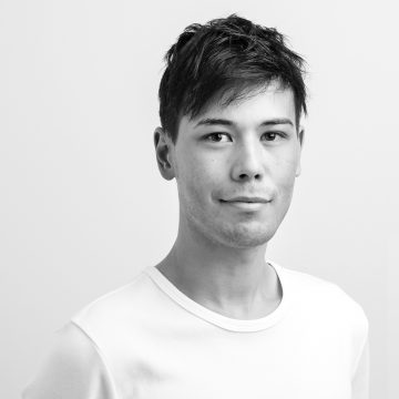 Nuori mies, jolla on lyhyet tummat hiukset ja jolla on valkoinen paita, poseeraa vaaleaa, yksinkertaista taustaa vasten. Hän katsoo suoraan kameraan hymyillen. Kuva on mustavalkoinen.