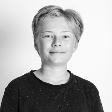 Mustavalkoinen muotokuva nuoresta henkilöstä, jolla on lyhyet, vaaleat hiukset, yllään tumma, pilkullinen t-paita, hymyilee hellästi ja katsoo kameraan vaaleaa taustaa vasten.