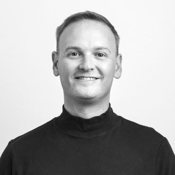 Hymyilevä mies, jolla on lyhyet hiukset ja korkea kaula-aukkoinen musta paita, kuvattu mustavalkoisena valkoista taustaa vasten.