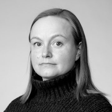 Naisella, jolla on suorat, olkapään pituiset hiukset ja vakava ilme, on yllään tumma, uurrettu villapaita. Kuva on mustavalkoinen ja taustaltaan tavallinen.