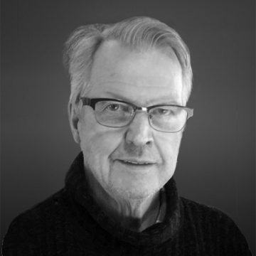 Muotokuva vanhemmasta miehestä, jolla on harmaat hiukset ja silmälasit, yllään tumma pusero, hymyilee hieman tumman taustan edessä.