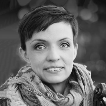 Lyhythiuksinen ja vaaleasilmäinen nainen, jolla on kuvioitu huivi, katsoo suoraan kameraan ja hymyilee kevyesti; tausta on pehmeästi sumea.
