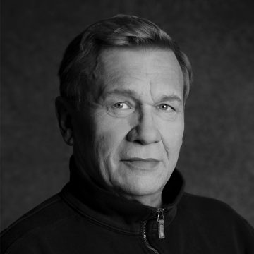 Keski-ikäinen mies, jolla on lyhyet vaaleat hiukset, katsoo kameraan neutraalilla ilmeellä. Hänellä on yllään tumma vetoketjullinen takki. Tausta on tavallinen ja tumma, ja kuva on mustavalkoinen.