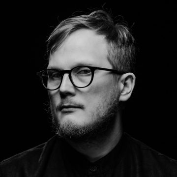 Mustavalkoinen muotokuva henkilöstä, jolla on lyhyet hiukset, silmälasit ja parta, yllään tumma paita, katselee hieman sivulle tummaa taustaa vasten.